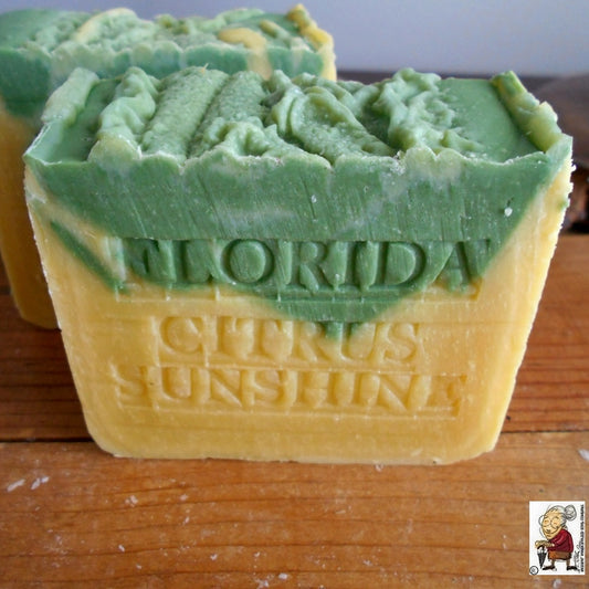 A Natural Citrus Handmade Soap Tangerine bar . The top layer is green with uneven, wavy textures, while the bottom layer is yellow. The Natural Citrus Handmade Soap Tangerine has the words "FLORIDA CITRUS SUNSHINE" embossed on it. There is a small logo in the bottom right corner from Natural Handcrafted Soap.